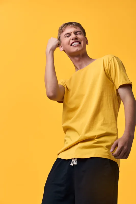 Man in a yellow shirt performs energetic dance movement against a bright studio backdrop, displaying a joyful pose, confident attitude and dynamic energy in a bold fashion moment