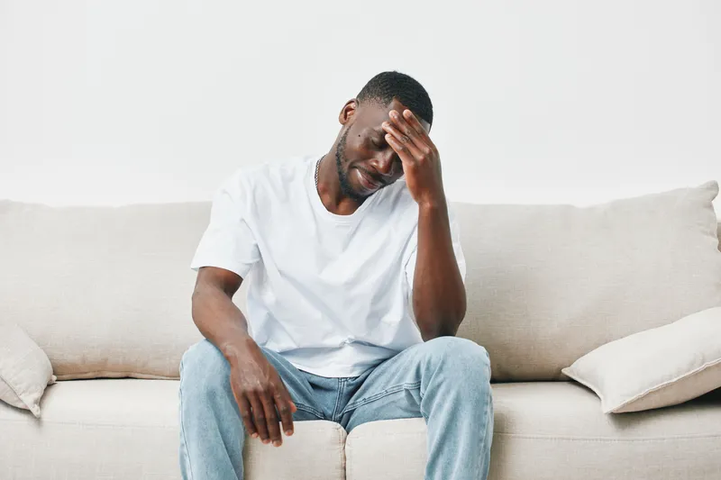 A young Black man in a white t shirt sitting on a beige sofa, showing signs of stress, capturing a moment of emotional struggle in a minimalist setting
