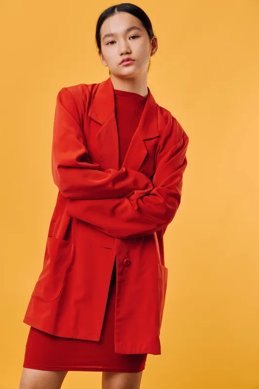 Woman wearing red blazer and dress standing with crossed arms against orange background. Young female model posing in fashion outfit with confident expression and casual style.