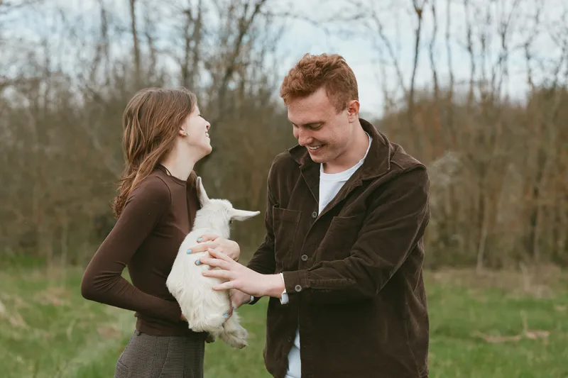 A couple in casual clothing gently cradle a goat kid in a sunny rural field, sharing a tender, playful moment. A candid lifestyle scene emphasizing care, nature, animals, and farm life.