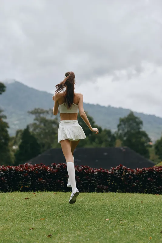 A young woman in a white athletic outfit running joyfully through a green lawn in a picturesque outdoor setting, reflecting a healthy lifestyle and vitality