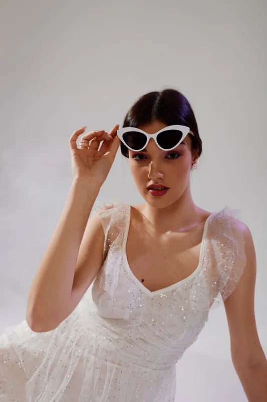 Young woman in a white dress wearing stylish sunglasses looks confidently at the camera. Soft lighting highlights her beauty and fashion sense in a studio setting.