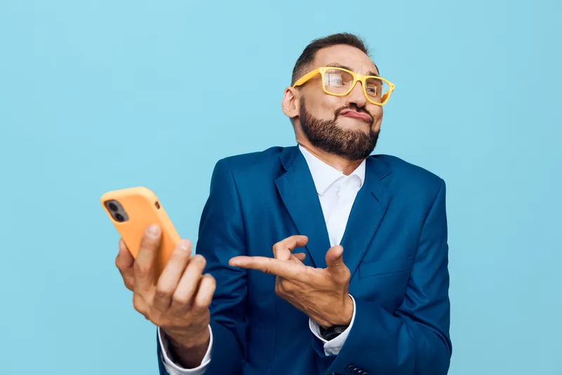 A confident business professional man in a blue suit stands against a solid colored background, using a smartphone and showing curiosity, focus, and a friendly, assertive mood through his gesture.