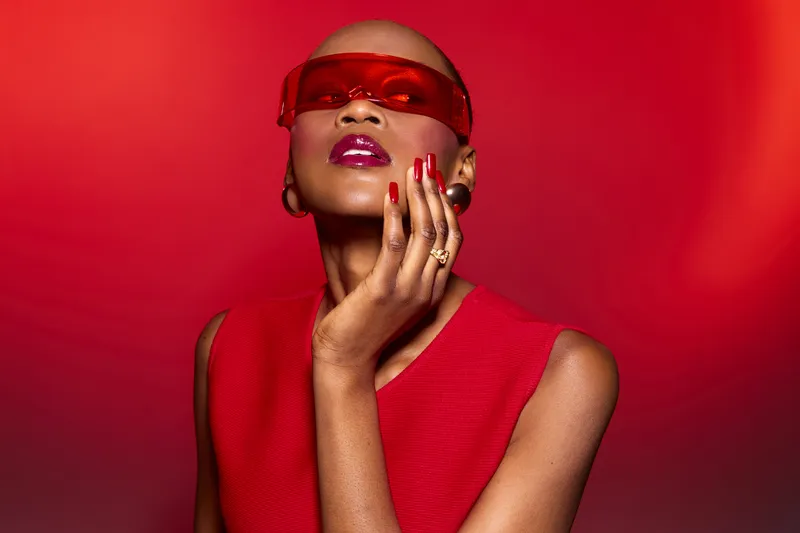 Stylish dark skinned African woman with shaved head wearing red sleeveless top and red sunglasses poses against red background. Concept of fashion, beauty, confidence, and modern style.