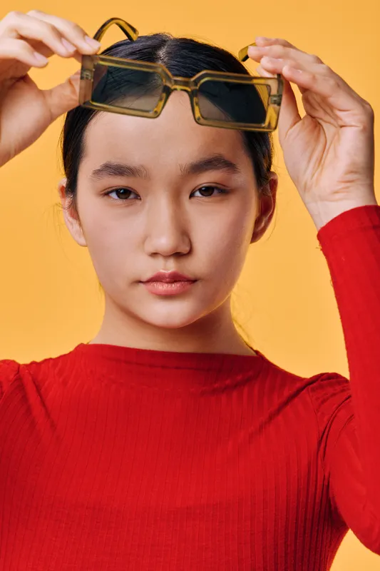 Young woman wearing red sweater and holding sunglasses above her head with serious expression on yellow background. Portrait of confident female with neat hairstyle indoors.
