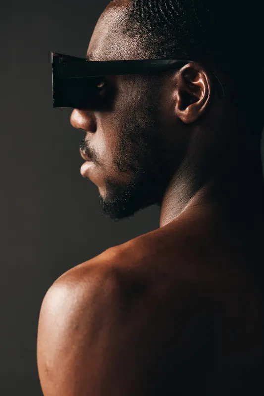 Creative conceptual portrait of African man with futuristic sunglasses and wet skin in studio. Dark background highlights visual style and modern artistic expression of male model.