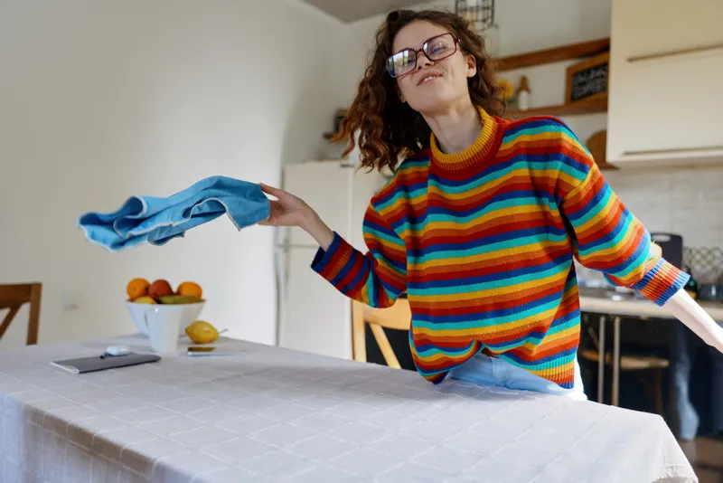 A woman in a colorful striped sweater irons a cloth on a kitchen table, capturing a casual home laundry moment, candid lifestyle scene focused on everyday chores and self expression.