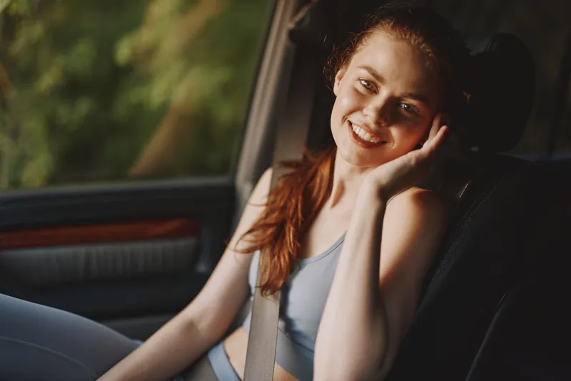 Cheerful young woman in sportswear sitting in a car, smiling, enjoying a sunny day, with sunlight filtering through trees, showcasing happiness and fitness