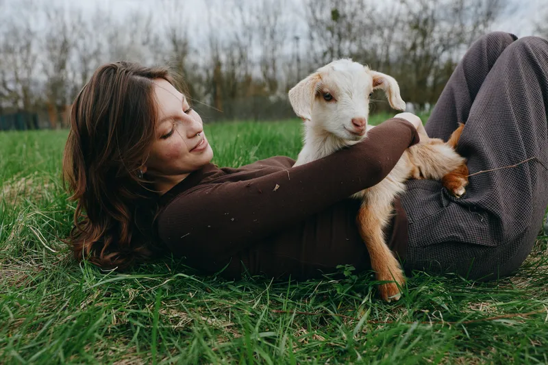 Goat, kid, woman, grass, outdoors, nature, meadow, field, animal, cuddle in a calm lifestyle shot showing a woman lying on grass while a goat kid rests in her arms, peaceful mood and gentle connection