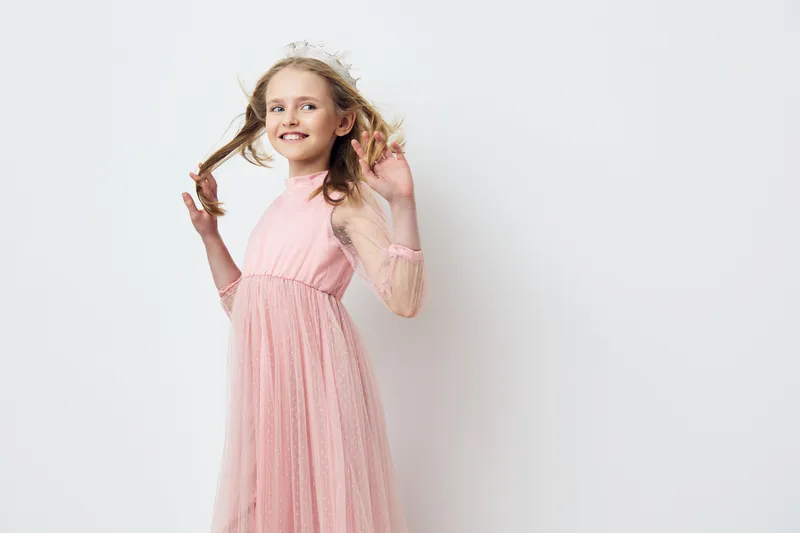 Cheerful young girl in a pink dress smiling and playing with her hair against a light background, perfect for themes of childhood and joy