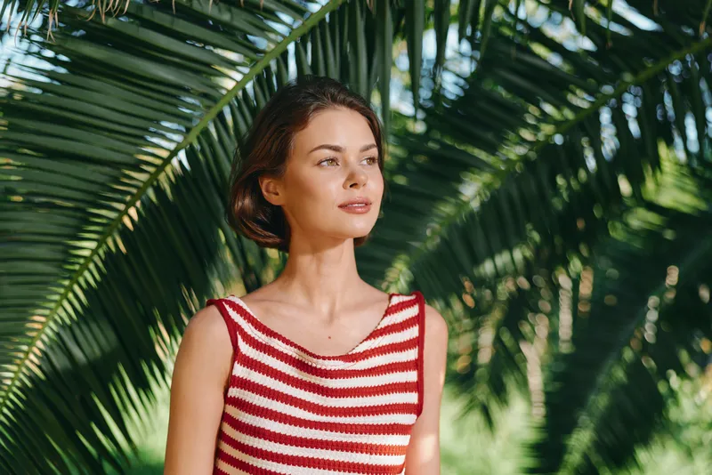 A stylish woman stands in a tropical garden wearing a red and white striped sleeveless top. She gazes into the distance, bathed in warm sunlight, with calm, confident presence.