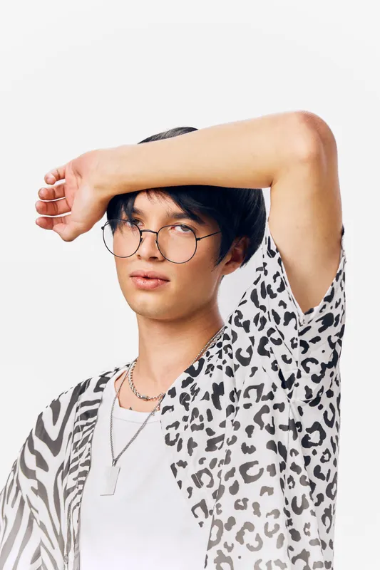 Young man wearing round glasses and animal print shirt posing with arm raised against white background, showcasing modern style and confident expression.