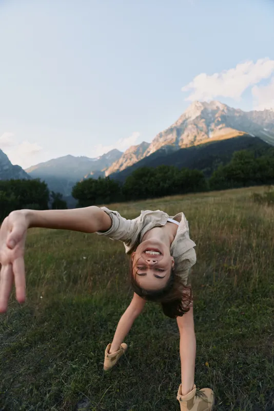 A smiling child performs a backbend in a meadow with mountains in the background, capturing carefree outdoor play, movement and balance in a bright nature scene.