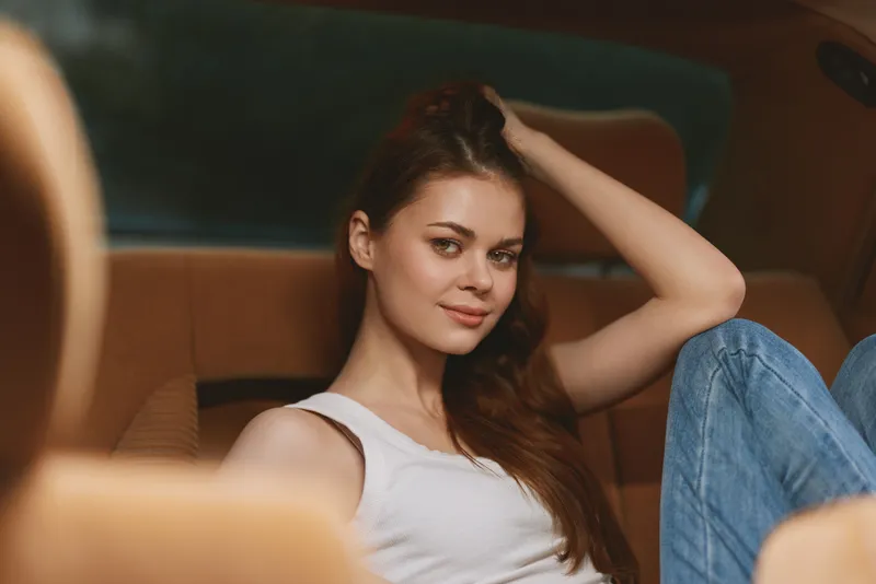 Young woman in casual attire relaxing in a car interior, showcasing a serene expression with natural light highlighting her features and soft brown tones.
