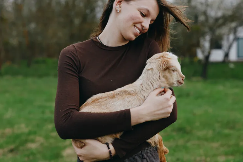 Joyful woman outdoors holds a young goat kid in her arms, smiling warmly as they share a tender moment in a green park, showcasing countryside lifestyle and a gentle bond.
