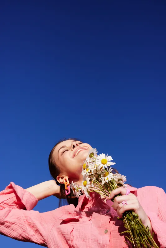 Young woman enjoying a sunny day with flowers, embodying happiness and warmth, in a vibrant pink outfit against a clear blue sky