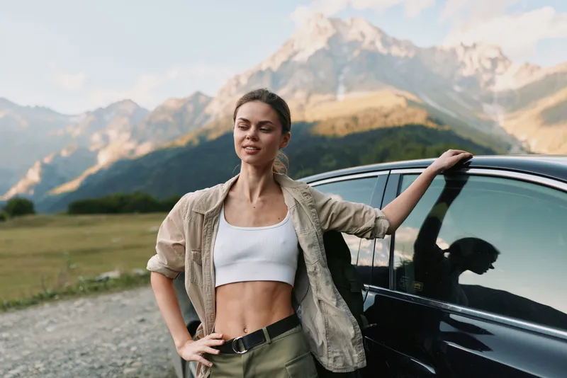 A confident young woman in casual outdoor clothing stands next to a black car with mountains in the background. The scene represents lifestyle and adventure.