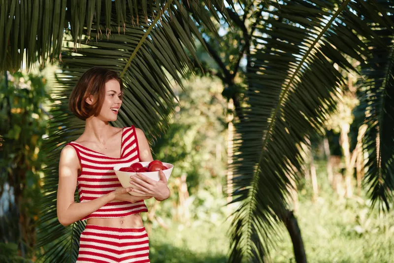 A cheerful woman in a red and white striped top stands in a lush tropical garden, smiling and holding a bowl of fruit. Sunlight filters through palm fronds, creating a bright, relaxed outdoor scene.