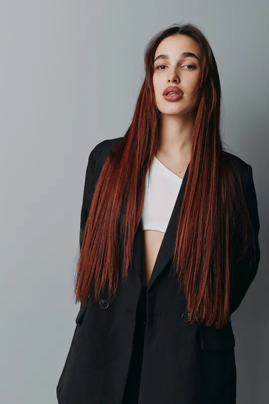 Woman with long red hair wearing black blazer and white top standing against gray background. Fashion model posing confidently with straight hair and neutral makeup for portrait photography.
