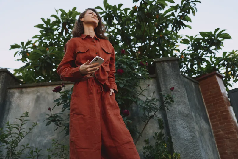 A young woman in a rust jumpsuit stands outdoors, looking up while holding a phone, against a garden wall, expressing curiosity and confident style