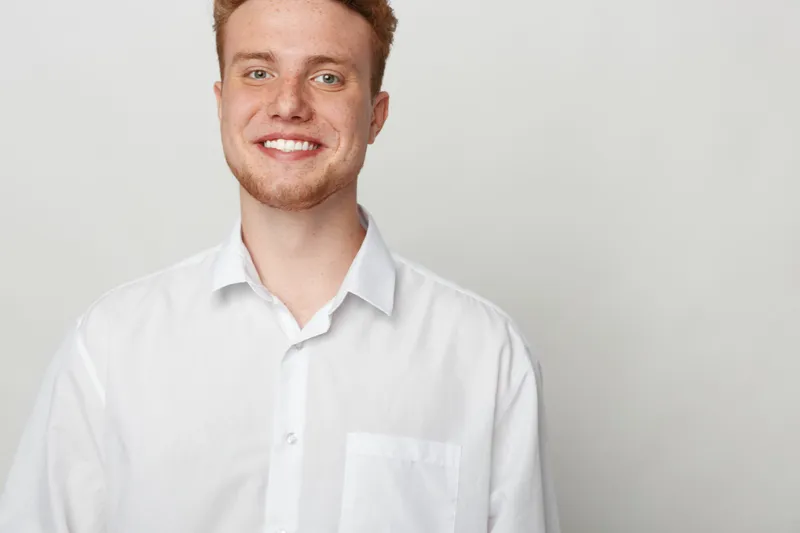 Smiling young man with red hair and beard wearing white shirt stands against plain light background. Portrait of confident male model with casual style and friendly expression.