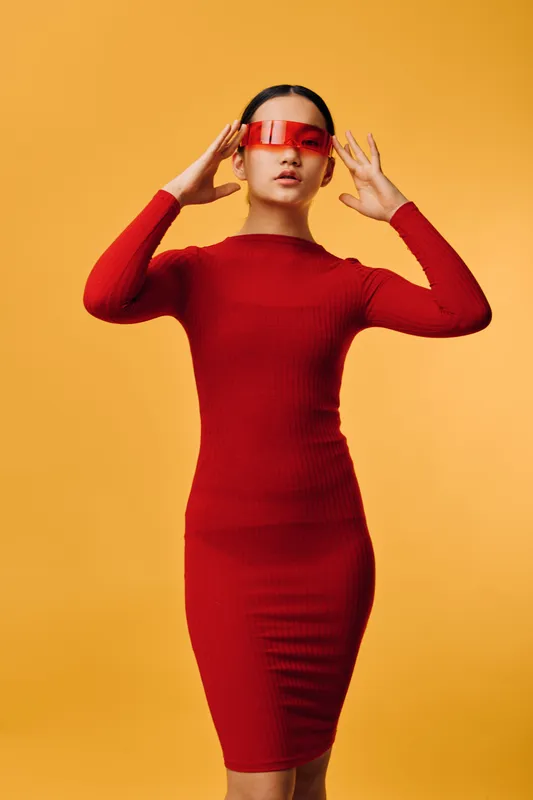 Woman wearing red fitted dress and red transparent glasses poses on yellow background with hands near head. Fashion style, modern look and confident female model in studio with bright lighting.