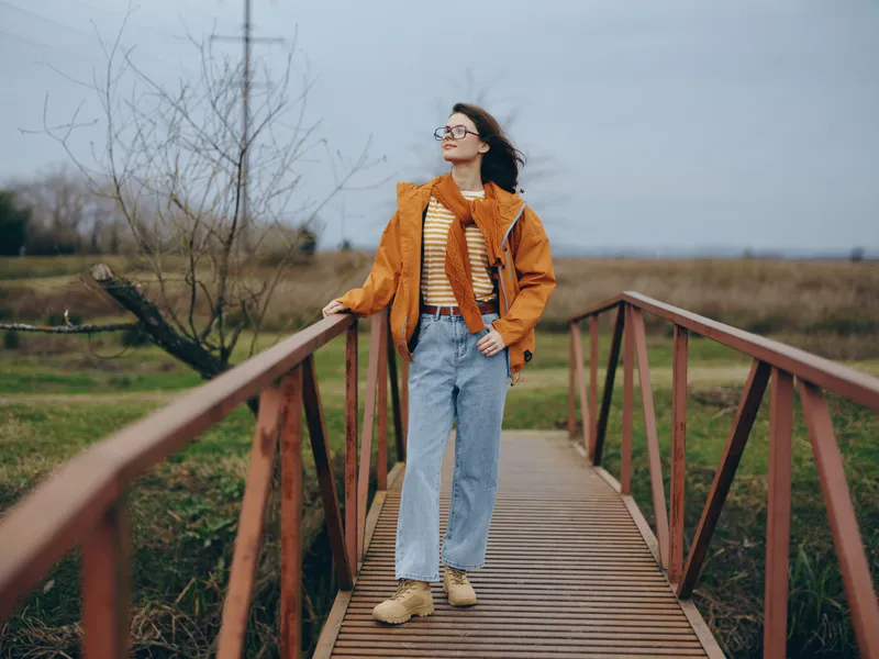 Woman lifestyle outdoors on a wooden bridge, wearing an orange jacket, denim jeans, glasses and scarf, standing in wind on a boardwalk over green field, cloudy sky, calm confident mood