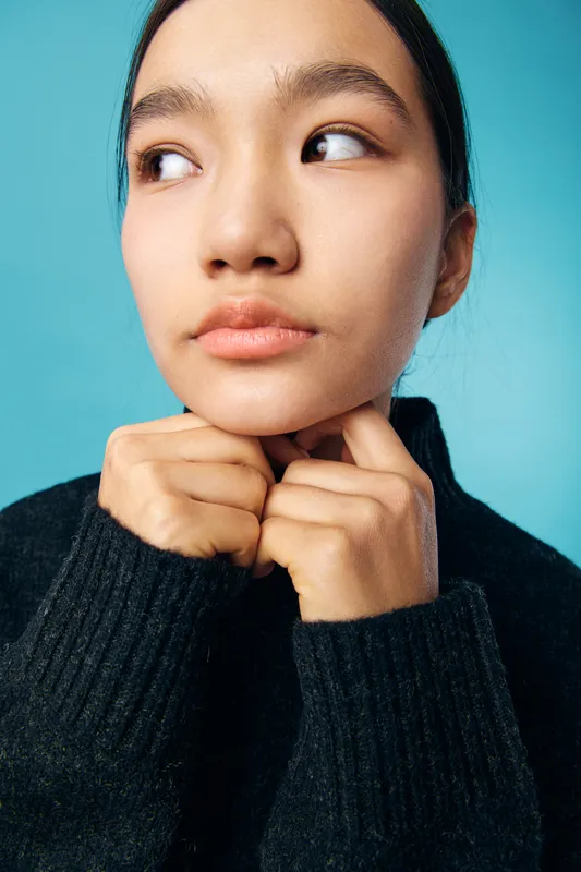 Young woman with clear skin looks thoughtfully aside, hands near chin, wearing black sweater. Closeup portrait on blue background with natural makeup and calm expression in studio lighting.