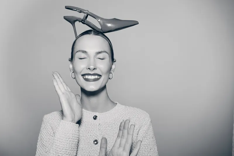 Happy white woman with fair skin and closed eyes balancing high heel shoe on head, smiling and posing in studio.