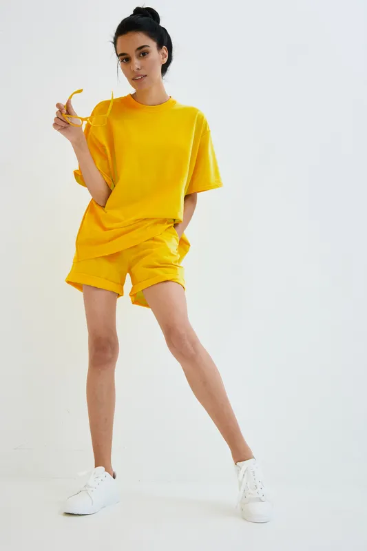 Young woman in bright yellow casual outfit poses confidently holding sunglasses with one hand, wearing white sneakers against clean white background for summer style.