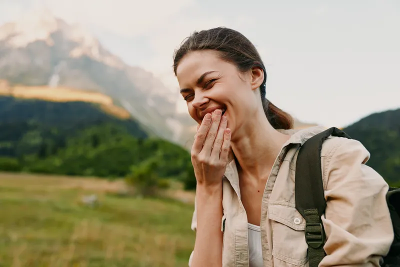 A joyful woman outdoors smiles with her hand near her mouth, wearing casual clothes and a backpack. A natural landscape behind her conveys spontaneous travel moments and a lighthearted mood.