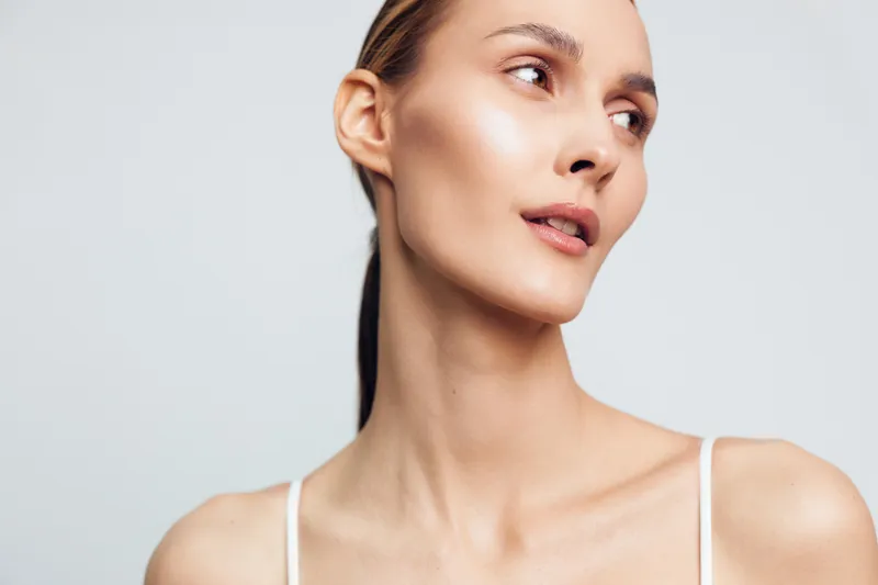 A closeup portrait of a young woman with clear skin and minimal makeup looking sideways against a plain light background. Concept for beauty, skincare, and natural health.
