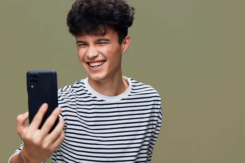 smiling man wearing striped shirt laughing joyfully while holding smartphone in hand, casual vibe with neutral background and natural lighting