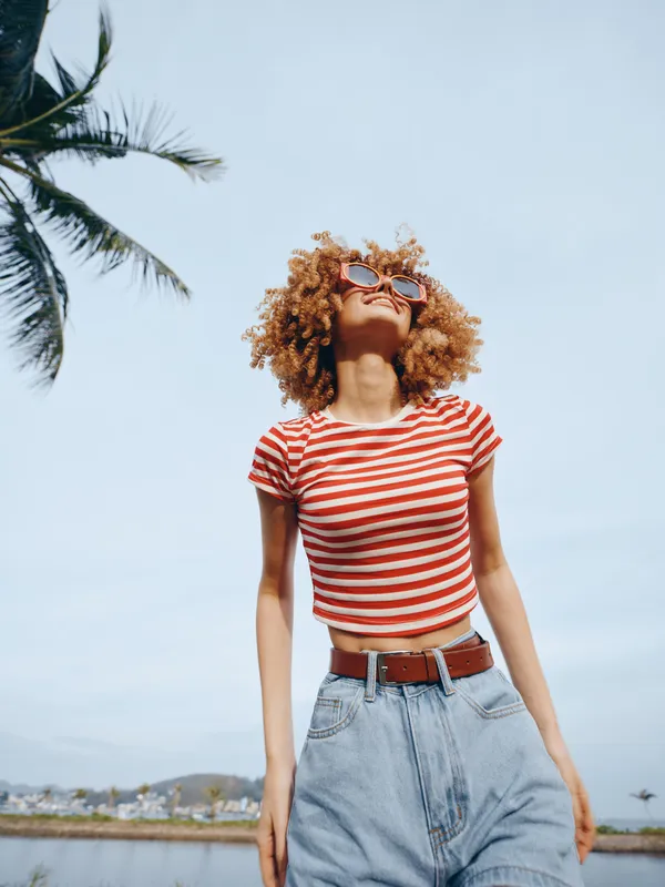 Portrait of a young woman with curly hair wearing a red and white striped crop top and high waisted denim jeans, standing confidently under a bright blue sky.