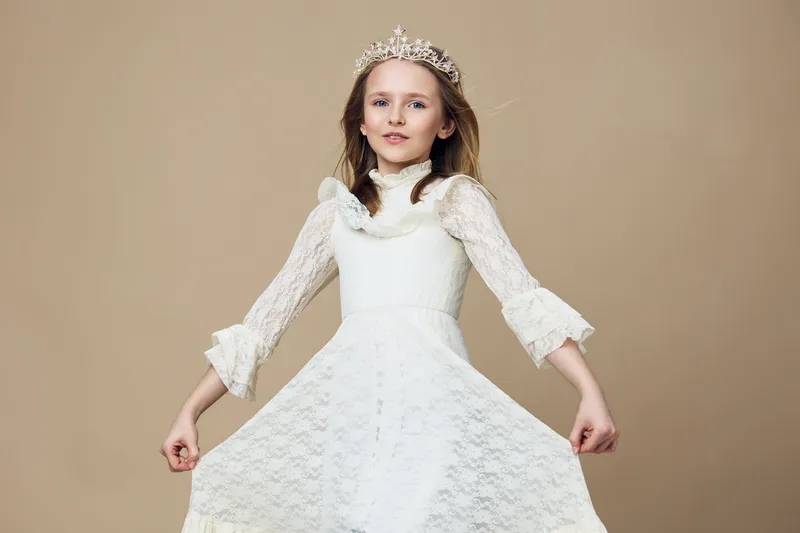 Elegant young girl in a white lace dress with a crown, showcasing confidence and joy against a neutral background, perfect for children s fashion and beauty themes