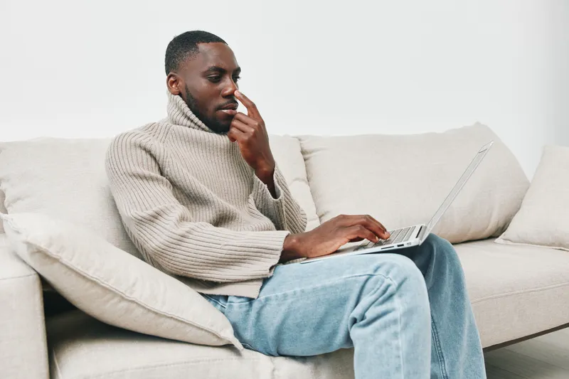 Young Black man in a cozy beige sweater sitting on a sofa, thoughtfully using a laptop in a modern, minimalist living room setting