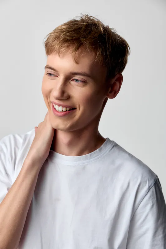 Smile, man in casual tshirt touches his neck in a relaxed pose with soft lighting, conveying warmth, approachability and everyday charm for lifestyle use