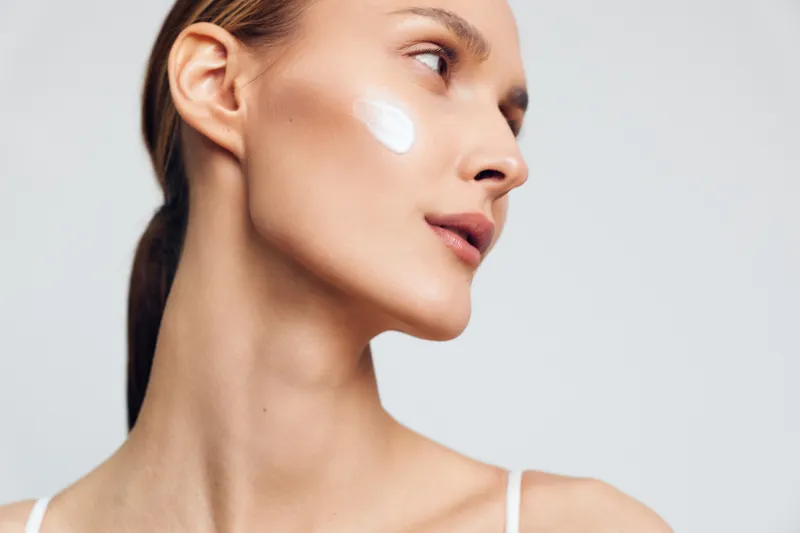 A young woman with clear skin applies white cream on her cheek against a plain light background. The image represents skincare, beauty, and daily facial care routine.