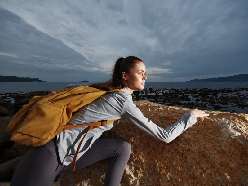 A young woman of Caucasian descent climbing a rocky surface by the sea, showcasing determination and adventure in a sporty outfit with a yellow backpack
