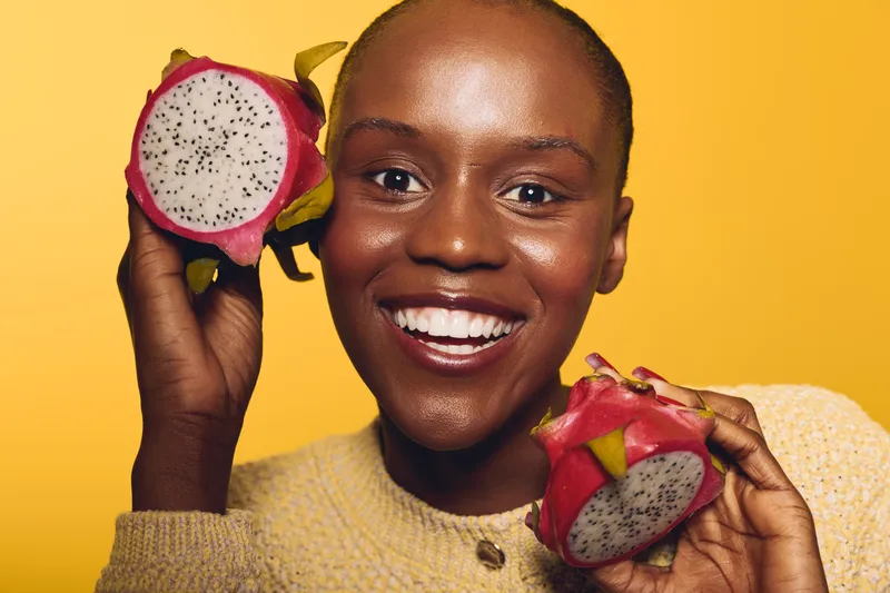 Happy African woman with dark skin holds two halves of a dragon fruit near her face, smiling brightly on yellow background. Concept of healthy lifestyle, exotic fruit, and joy.