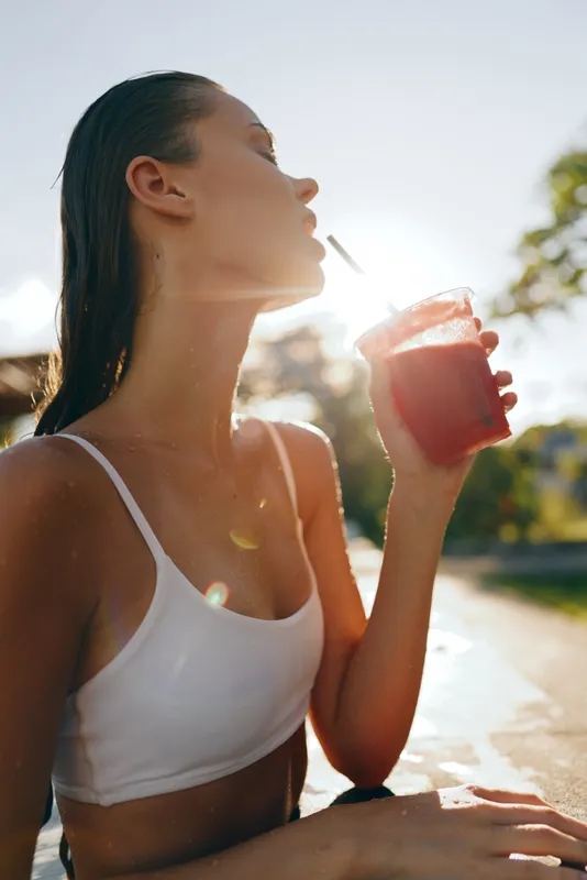 Cheerful woman outdoors, sipping juice under warm sun, casual fitness vibe, relaxed posture, soft breeze, natural colors, vibrant background, everyday lifestyle moment.
