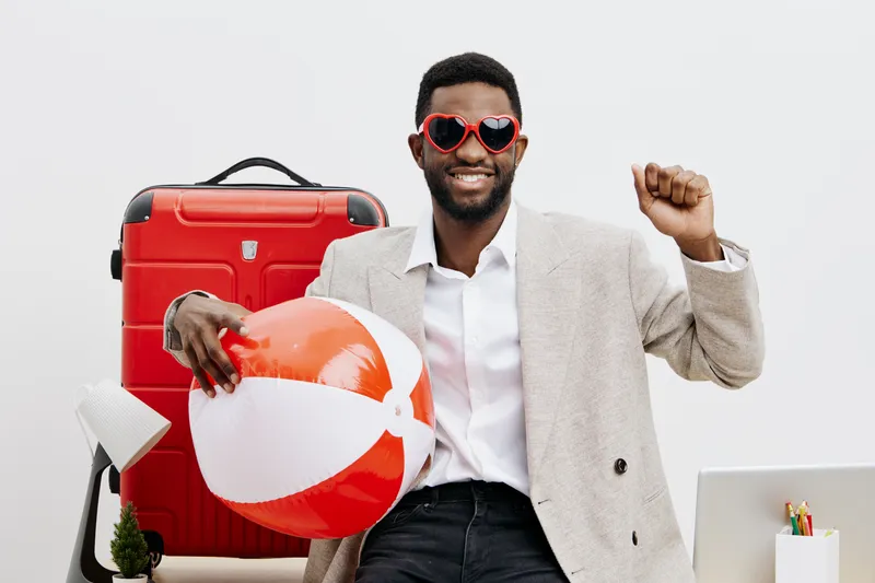 Smiling man with dark skin holding an orange beach ball, standing in front of a red suitcase, with a cheerful expression on his face, dressed in a light blazer and white shirt, travel concept, summer 