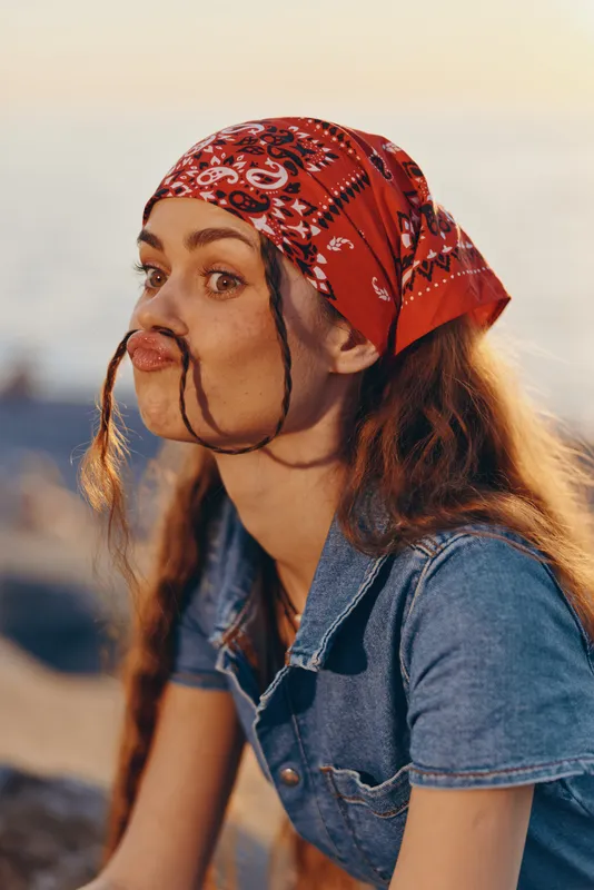 Young woman wearing red bandana and denim shirt with braided hair sits outdoors with sunset light and calm background, enjoying a peaceful moment.