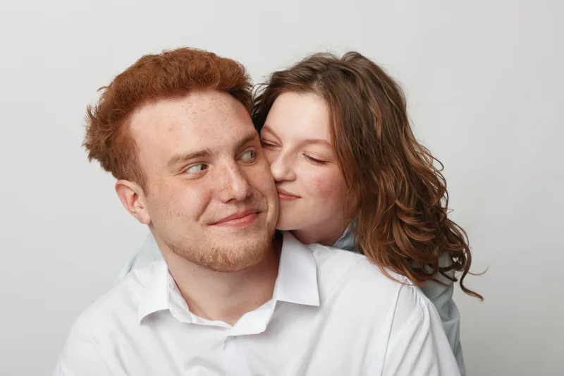 Young couple with red hair and curly hair share a tender moment, woman hugging man from behind. Intimate love, affection, and happiness in a bright studio setting.