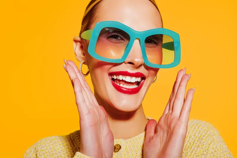 Smiling woman with bright red lipstick wears large turquoise sunglasses and yellow sweater on vibrant orange background. Concept of summer fashion, happiness, and colorful style.