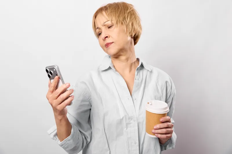 elderly woman indoors holding smartphone and coffee cup, tired worried expression, blonde short hair, white studio background, casual shirt, soft even lighting, waiting moment