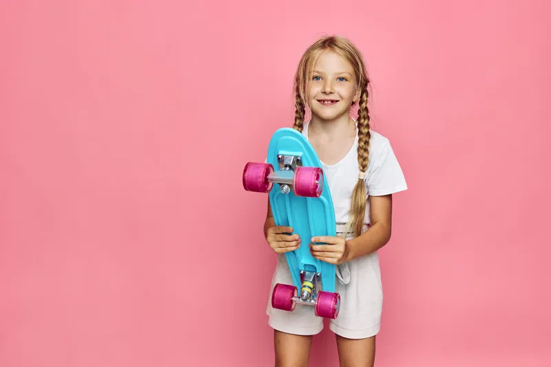 Smiling girl with blonde braids holding a blue skateboard with pink wheels on a pink background.