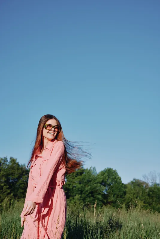 Smiling young woman in a pink dress enjoying a sunny day outdoors with flowing hair and a clear blue sky, evoking joy and freedom in nature