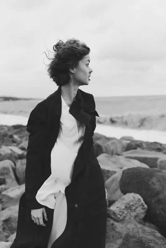 Woman in a long coat stands on a windswept coastline, gazing toward the ocean among rugged rocks, captured in monochrome for moody fashion storytelling
