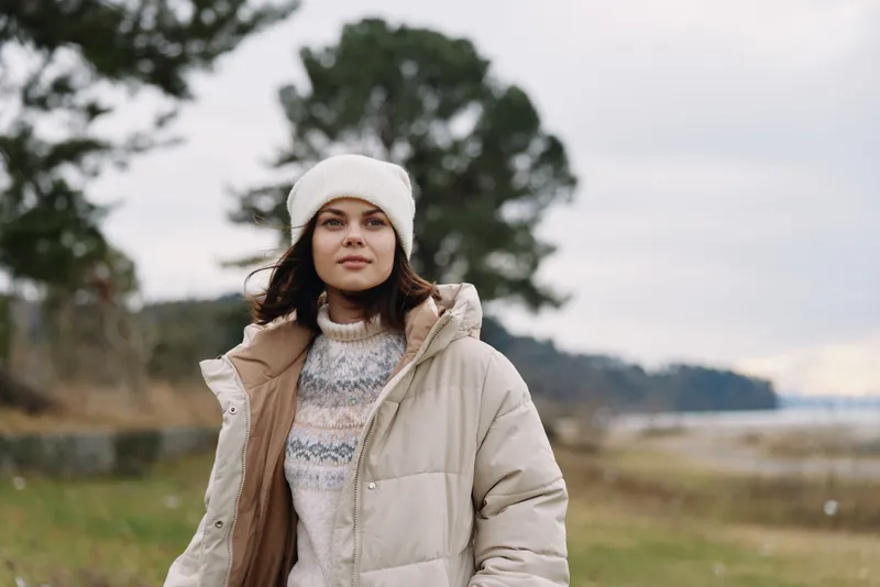 Outdoor portrait of a woman in a warm coat and knit sweater by a windy shoreline, with trees and calm water, showcasing winter fashion and serene nature